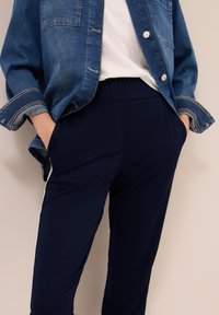 Persona che indossa pantaloni jogger blu scuro con elastico in vita, striscia laterale bianca, t-shirt bianca e giacca di jeans blu aperta con maniche arrotolate.