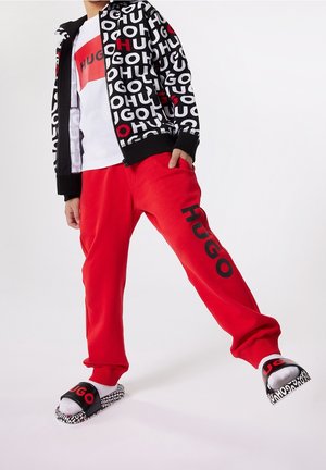 Bambino che indossa una giacca con motivo a lettere bianca e nera, camicia bianca, pantaloni rossi con la scritta "HUGO" e ciabatte nere con calzini bianchi.