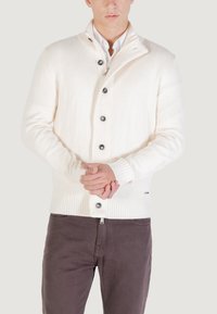Hombre vistiendo un cárdigan blanco abotonado sobre una camisa blanca y pantalones marrón oscuro, con las manos entrelazadas al frente contra un fondo claro.