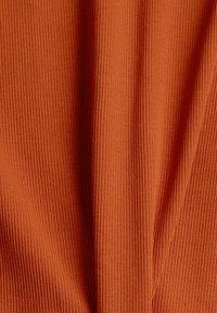 Tissu côtelé de couleur terre cuite chaude, présentant une surface texturée avec des lignes verticales créant une profondeur et une dimension subtiles.
