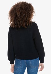 Suéter de punto negro con patrón texturizado, corte holgado y puños y dobladillo acanalados. Combinado con jeans azules, mostrando la vista trasera.