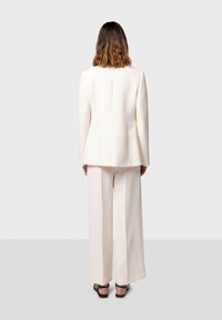 Blazer bianco sartoriale e pantaloni a gamba larga. Tessuto liscio con dettagli di cucitura minimi. Vista posteriore aperta, abbinato a sandali neri.