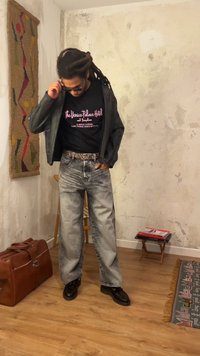 Άνδρας με dreadlocks διορθώνει τα γυαλιά ηλίου, φοράει μαύρο δερμάτινο μπουφάν, μπλουζάκι με τύπωμα, τζιν με acid wash και μαύρα παπούτσια σε ένα ρουστίκ δωμάτιο.