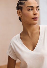 Weißes Baumwoll-T-Shirt mit V-Ausschnitt und kurzen Ärmeln. Das Modell hat geflochtenes Haar und trägt kleine Ohrstecker; neutraler Hintergrund mit weichem Licht.