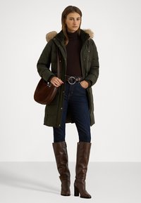 Cappotto imbottito verde con cappuccio foderato in pelliccia, indossato sopra un dolcevita scuro e jeans blu, abbinato a stivali alti marroni con tacco e una borsa marrone.