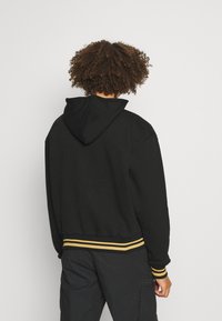 Sudadera negra con capucha ajustable, puños acanalados y dobladillo con rayas amarillas y negras. Tejido suave y texturizado, corte relajado.