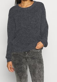 Mörkgrå stickad tröja med rund halsringning och långa ärmar, kombinerad med gråa figurnära jeans, som visar upp ett texturerat tyg.