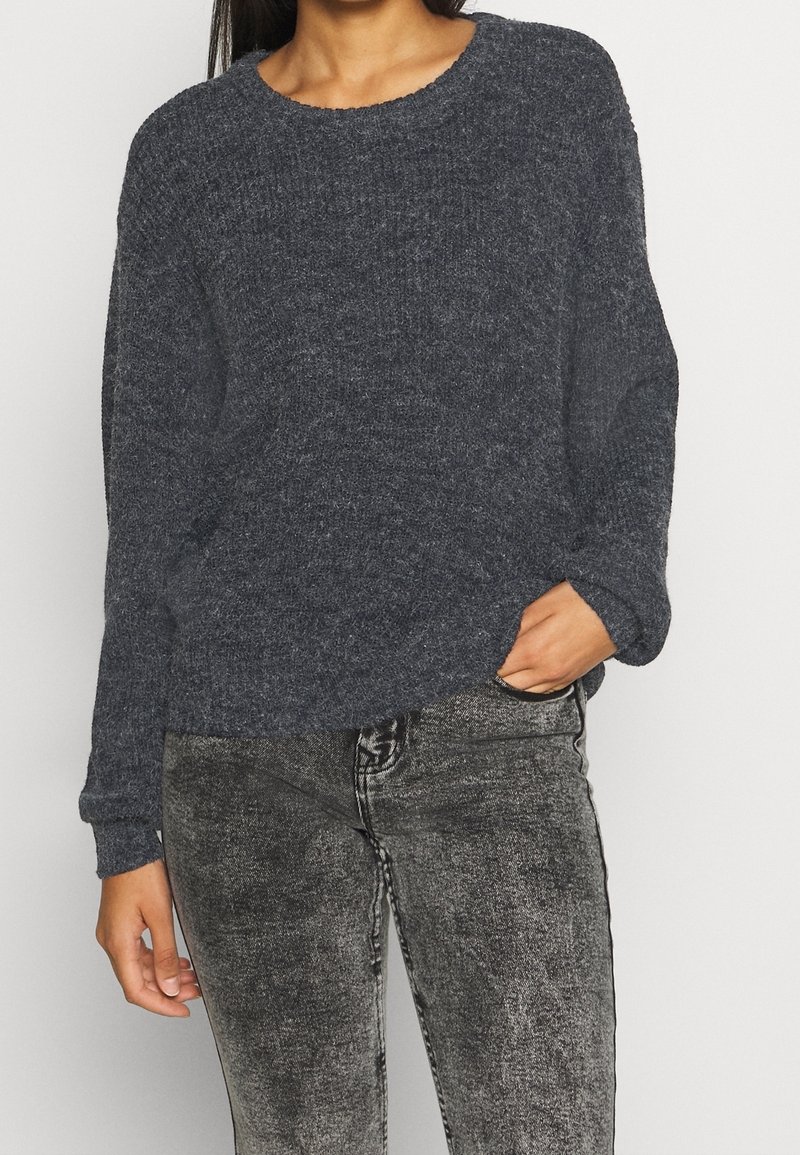 Mörkgrå stickad tröja med rund halsringning och långa ärmar, kombinerad med gråa figurnära jeans, som visar upp ett texturerat tyg.