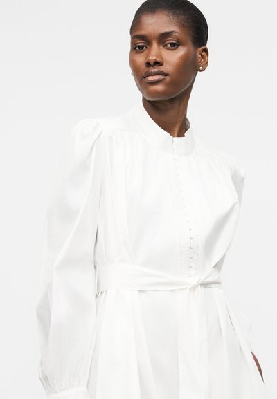 Γυναίκα με σκούρο δέρμα που φοράει λευκό φόρεμα με ψηλό λαιμό, μακριά φουσκωτά μανίκια και δεμένη ζώνη στη μέση, ποζάρει μπροστά σε απλό φόντο.