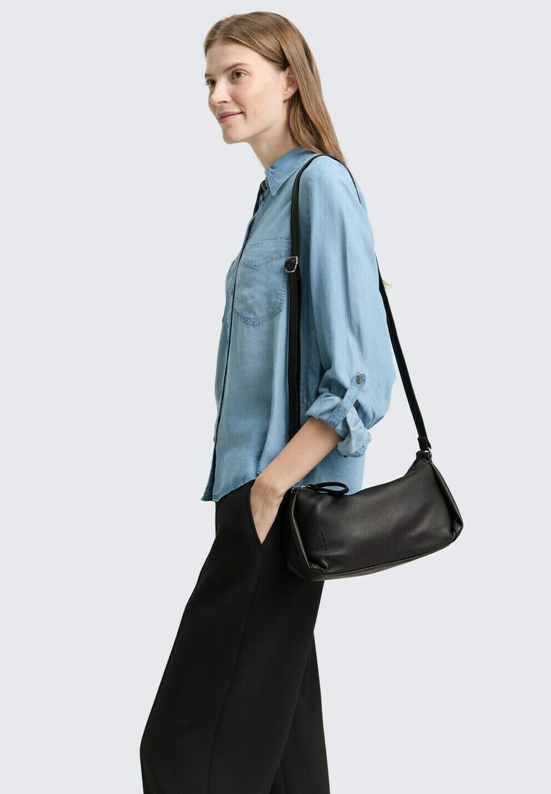 Borsa a spalla in pelle nera con una superficie liscia, forma curva e tracolla rimovibile, indossata crossbody su uno sfondo azzurro chiaro.