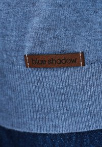 Jasnoniebieska dzianina ma wyrazisty wzór. Brązowa skórzana metka z białym przeszyciem nosi napis "blue shadow".