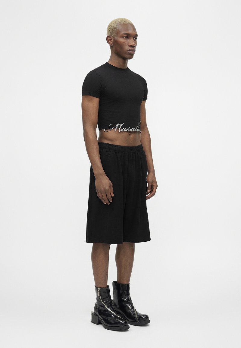 Man with blond hair wearing black cropped t-shirt with text, knee-length black shorts, and shiny black ankle boots, standing against plain background.