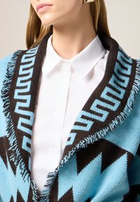 Scialle di lana con un motivo a zigzag in turchese e marrone, con bordi frangiati. Abbinato a una camicia bianca con colletto.