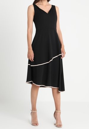 Sort ærmeløs kjole med V-hals, der har en lagdelt nederdel med cremefarvet kant. Fremstillet af et glat, flydende stof.