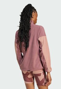 Fleece-takki mauvessa, jossa on kontrastoivat vaaleanpunaiset hihat; mukana korkea kaulus ja pehmeä rakenne. Yhdistetty kuvioituihin pyöräilyshortseihin.