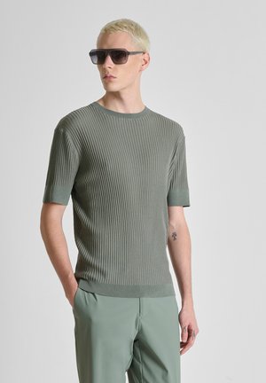 Hombre con cabello rubio corto que lleva gafas de sol oscuras, suéter verde de manga corta acanalado y pantalones verdes, de pie con las manos en los bolsillos.