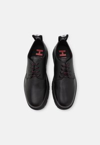 Une paire de chaussures noires en cuir avec lacets, dotées de semelles épaisses et de lacets accentués de rouge, vue de dessus sur un fond blanc.