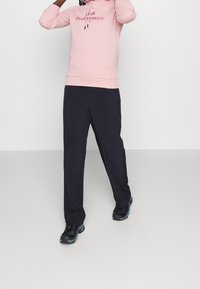 Felpa rosa chiaro con tasca frontale e cordino, abbinata a pantaloni blu scuro e scarpe da ginnastica nere. Abbigliamento sportivo semplice e casual.