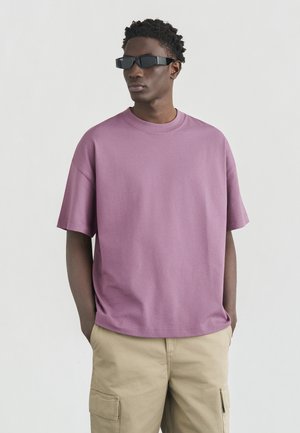 Uomo che indossa occhiali da sole rettangolari neri, t-shirt viola larga e pantaloni cargo beige, in piedi con le mani nelle tasche davanti a uno sfondo semplice.