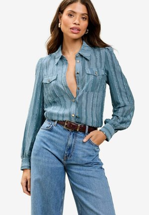 Blauw gestreepte blouse met een v-hals, twee borstzakken en knoopdetails. Gecombineerd met high-waisted blauwe jeans en een bruine riem.