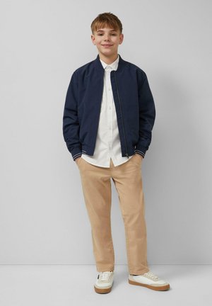 Garçon debout les mains dans les poches, portant une veste bomber bleu marine, une chemise blanche boutonnée, un pantalon beige et des baskets blanches sur un fond gris.