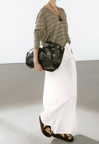 Sac en cuir noir avec des œillets circulaires, texture douce, forme asymétrique. Le mannequin porte un haut beige rayé et un pantalon blanc à jambes larges.