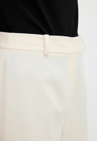 Beige broek van een gladde stof, met een vlakke tailleband en ceintuurlussen. De stof lijkt lichtgewicht en heeft een licht glanzende afwerking.