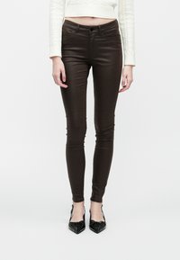 Hnedé úzke džínsy majú hladkú textúru, stredne vysoko posadený pás, päť vreciek a priliehavý strih, skombinované s čiernymi lodičkami s ostrou špičkou.