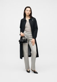 Γυναίκα που φοράει μακρύ μαύρο παλτό, μπεζ τοπ, ανοιχτό γκρι τζιν, μαύρες γόβες και κρατά μια μικρή μαύρη τσάντα, πάνω σε μονόχρωμο φόντο.