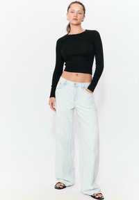 Schwarzes, eng anliegendes Langarmoberteil kombiniert mit hellblauen, weiten Jeans mit Knopfverschluss an der Taille und seitlichen Taschen, getragen mit schwarzen Sandalen.