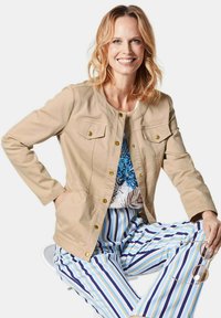 Beige, aansluitende jas met gouden knopen en twee borstzakken. Gecombineerd met gestreepte blauw-witte broek. Model houdt een zonnebril vast.