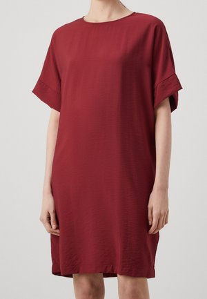 Femme portant une robe ample rouge foncé à manches courtes, arrivant au genou, avec un col rond, sur un fond clair uni.