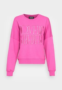 Růžová mikina vyrobená z měkké tkaniny s kulatým výstřihem, dlouhými rukávy a žebrovanými manžetami. Na přední straně má výrazné, texturované písmo.