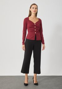 Burgundy hosszú ujjú blúz négyzet alakú nyakkivágással, ráncolt részletekkel, öt aranyszínű gombbal, fekete bő szárú nadrággal és hegyes orrú magassarkú cipővel párosítva.