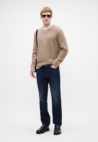 Beige textúrájú, V-nyakú pulóver, sötétkék egyenes szabású farmernadrág és fekete cipő. A modellen napszemüveg van, és egy fekete válltáska van nála.