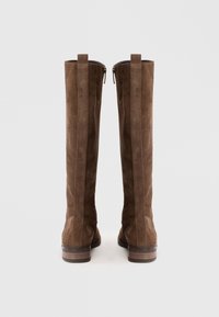 Botas altas de ante marrón con una textura suave, cierre de cremallera en la parte posterior y un tacón de madera apilada. El diseño presenta costuras verticales sutiles.