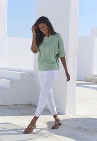 Helles grünes, strukturiertes Oberteil mit kurzen Ärmeln, kombiniert mit weißen, taillierten Hosen mit seitlichen Reißverschlüssen und braunen Sandalen mit dekorativen Akzenten.