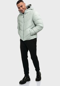 Hellgrüne gepolsterte Jacke mit Kapuze, diagonal genäht und mit Reißverschluss, kombiniert mit schwarzen Hosen und robusten schwarzen Stiefeln.