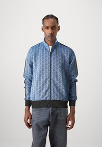 Blå zip-up jakke med et geometrisk mønster, sort ribbet manchet og kant, samt kontrasterende sidelister. Lacoste-logoet er vist på brystet.