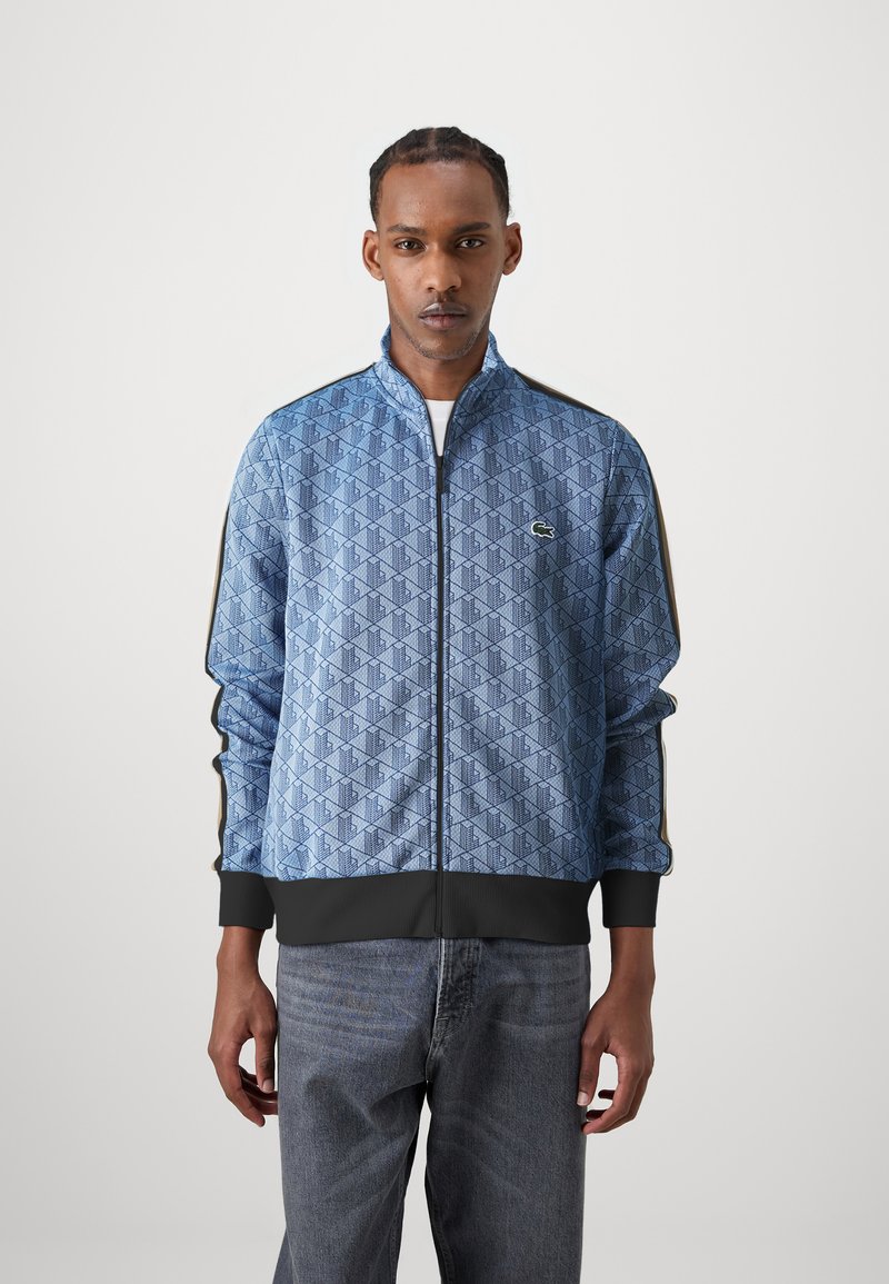 Blå zip-up jakke med et geometrisk mønster, sort ribbet manchet og kant, samt kontrasterende sidelister. Lacoste-logoet er vist på brystet.