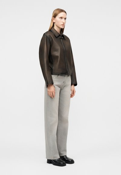 Γυναίκα στέκεται φορώντας σκούρο καφέ δερμάτινο μπουφάν, ανοιχτό γκρι παντελόνι με φαρδύ πόδι και μαύρα παπούτσια με τετράγωνη μύτη σε απλό φόντο.