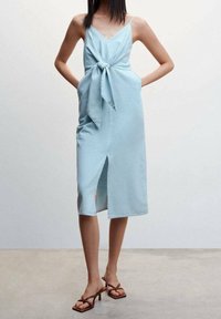 Sininen farkkumekko, jossa on solmittava etuosa, V-aukko, sivutaskut ja reiteen asti korkea halkio. Sujuva rakenne, polven puoliväliin asti ulottuva pituus.