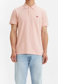 Mężczyzna w jasnoróżowej koszulce z kołnierzykiem polo z czerwonym logo Levi's oraz jasnoniebieskich dżinsach, stojący na tle białym.