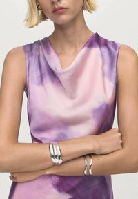Sukienka bez rękawów w fioletowo-różowy wzór akwarelowy, wykonana z gładkiego materiału. Posiada dekolt w drapowaniu. Na ramionach noszone są srebrne bransoletki.