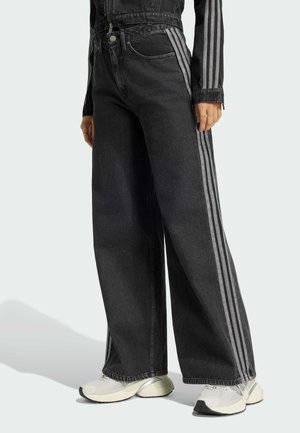 Person iført sorte jeans med vide ben med tre grå sidelister og hvide sneakers, der står mod en ensfarvet baggrund.