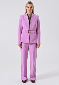 Vrouw die een lila blazer met ceintuur en bijpassende wijde broek draagt over een metallic top en donkere puntige schoenen tegen een grijze achtergrond.