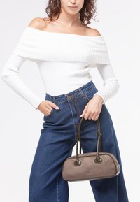 Donna che indossa un top bianco a spalle scoperte a coste e jeans blu a vita alta, mentre tiene in mano una borsa marrone con doppi manici.