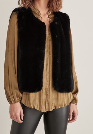 Femme portant un gilet en fausse fourrure noire sur un chemisier plissé marron et un pantalon en cuir noir, debout devant un fond uni.