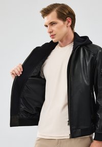 Zwarte leren jas met een ritssluiting aan de voorkant, voorzien van een capuchon en geribbelde manchetten. De binnenvoering is van glad zwart materiaal. Draag het over een lichtgekleurde shirt.