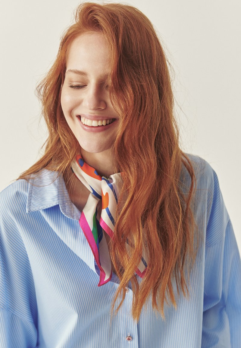 Svetlo modra srajca z belim ovratnikom, kombinirana s pisano svileno rutko z vzorcem, vezano okoli vratu. Valoviti rdeči lasje.